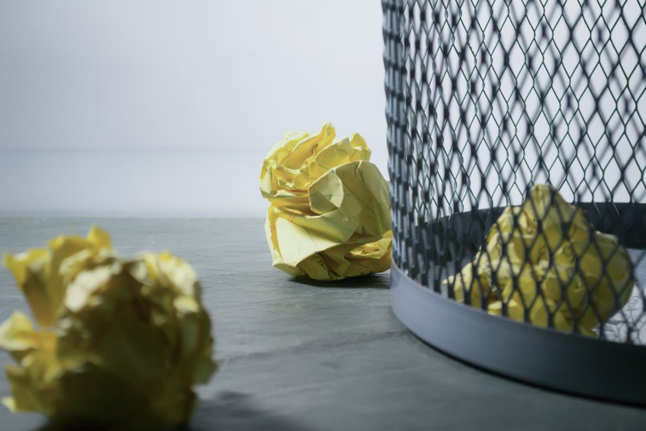 The image shows a small collection of crumpled yellow paper balls on a flat, smooth surface, likely a tabletop or floor, with three of them positioned towards the center and foreground. To the right, there is a cylindrical metal wire mesh bin with a rounded base, which is partially visible and contains additional yellow paper balls, indicating that the paper debris has been disposed of or is in the process of being discarded. The background features a plain white wall, providing a neutral backdrop that emphasizes the contrast between the yellow paper and the metallic bin. The lighting appears to be natural or softly diffused, highlighting the crumpled textures of the paper balls and the reflective surface of the wire bin. The scene subtly evokes themes of waste disposal and material recycling, aligning with the context of independent rubbish removal or designated disposal services, as offered by Rubbish Removal Dulwich, within an indoor setting focused on refuse management practices.