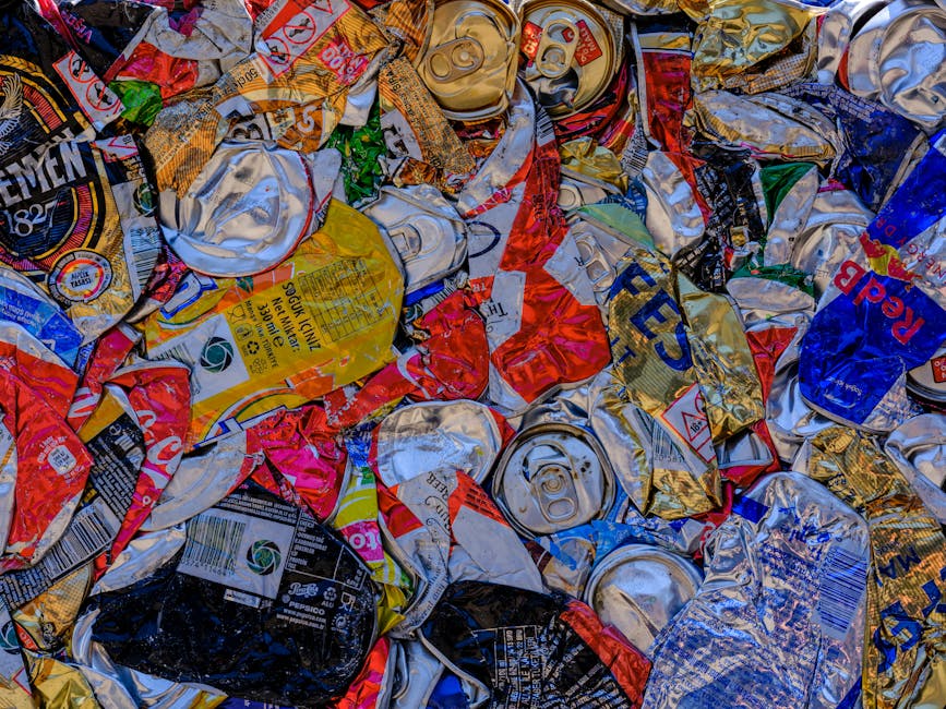 A close-up view of a large pile of crushed and crumpled aluminum cans and food tins, scattered haphazardly over a flat surface. The cans display a variety of colours, including silver, gold, red, blue, black, and yellow, with some featuring brand labels, logos, and nutritional information that are partially visible but distorted by the crumpling process. The metallic finishes reflect light, creating shiny highlights and shadows that emphasize their irregular shapes. Many cans have pull-tabs, some still attached, and the textures range from smooth and glossy to dented and scratched. The background shows an indistinct surface, possibly a pavement or a container floor, with no other objects or environmental features in view. This image is representative of the sort of recyclable metal waste collected separately for disposal or recycling, a process often undertaken by waste removal services such as Rubbish Removal Dulwich to promote environmentally responsible rubbish removal and alternative waste handling methods.