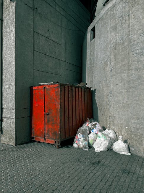 A red, industrial metal rubbish skip with a weathered and slightly corroded surface, positioned against a dark green tiled wall on the left and a light grey concrete wall on the right, situated outdoors on a paved area with dark grey bricks. Several white plastic rubbish bags filled with waste, some appearing partially torn or bulging, are stacked beside the skip, leaning slightly to the right. The scene is lit evenly with natural daylight, and the surrounding environment includes urban building facades, suggesting a private or non-municipal rubbish collection point used for site clearance or independent waste disposal services. The overall setting reflects a typical waste management scenario where bulky refuse is temporarily stored prior to collection, aligning with non-public, on-site rubbish handling practices occasionally employed by companies like Rubbish Removal Dulwich.