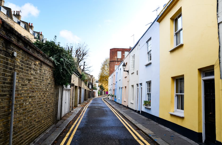 A narrow residential street paved with dark, wet asphalt, featuring a double yellow line along the curb. On the right, there are brightly painted terraced houses with smooth, pastel-colored facades in yellow, white, and pale blue, each with rectangular white-framed sash windows, some with small flower boxes below. The houses have simple, flat fronts with minimal decorative details. On the left, a weathered brick wall runs along the sidewalk, topped with greenery and small trees. In the background, a taller red-brick building with multiple stories and a flat roof is visible, partially obscured by trees with autumn foliage. The sky is partly cloudy with patches of blue, and the scene is evenly lit, suggesting it is daytime, possibly after a recent rain. The scene provides an example of a typical quiet street that could benefit from private rubbish removal services by a company like Rubbish Removal Dulwich, emphasizing the residential environment and narrow street conditions suited for on-site clearance.