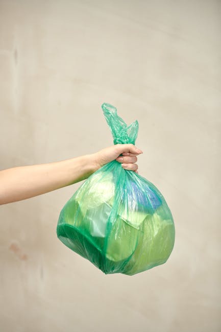 A person's hand is visible holding a green plastic rubbish bag with a twist knot at the top, against a plain, light-colored background. The bag appears to be filled with waste and is slightly translucent, showing some contents inside. The skin tone of the hand is light, and the arm is extending from the left side of the image. The environment suggests an indoor setting, potentially related to waste disposal, as part of a rubbish removal process. The image emphasizes individual, private waste handling, aligning with services like independent rubbish collection or on-site clearance offered by companies such as Rubbish Removal Dulwich. The focus is on the plastic bag, its texture, color, and the act of holding it ready for disposal, reflecting themes associated with rubbish removal and waste management services.