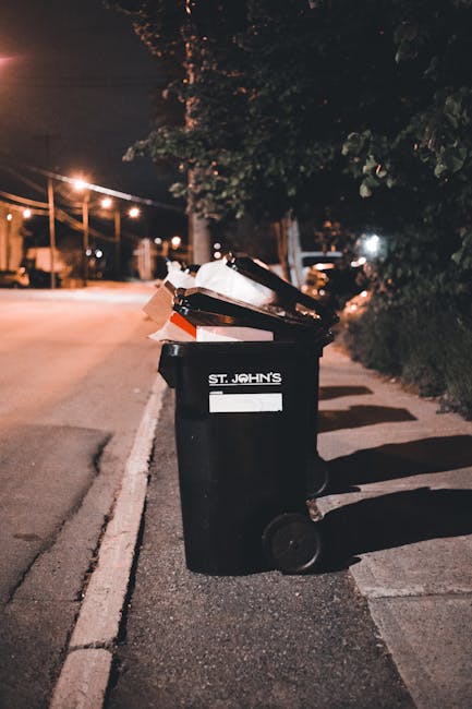 A black wheeled rubbish bin with a white label and the name 'St. John's' printed on the side is positioned on the pavement near the curb in an outdoor urban setting during the evening. The bin is open at the top and filled with various discarded items, including cardboard and plastic waste. Surrounding the bin is a concrete sidewalk with a curb, and the bin casts a shadow on the pavement due to nearby streetlights. Behind the bin, there are trees with dense foliage, and further back, a row of streetlights illuminates the street, with glimpses of parked cars and buildings visible in the distance. The scene conveys an environment where independent rubbish collection or on-site waste disposal services, such as those offered by Rubbish Removal Dulwich, operate to manage local waste efficiently outside residential or commercial properties.
