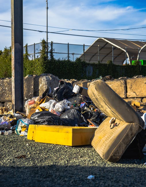 A pile of discarded rubbish placed on a gravel surface next to a stone wall, including black plastic bags overflowing with waste, a yellow plastic container, and a large, worn, beige upholstered armchair with fabric covering that appears soiled and slightly disintegrated. The background features a metal fence with greenery beyond, such as small trees or bushes, and an outdoor shelter or canopy structure with a curved roof. Overhead, there are power lines against a bright blue sky with some wispy clouds. The scene is illuminated by natural daylight, emphasizing the contrast between the waste materials and the surrounding environment. This image relates to private rubbish removal or alternative waste handling services, possibly illustrating unmanaged waste or the need for professional clearance, as seen in the context of rubbish removal services in Dulwich.
