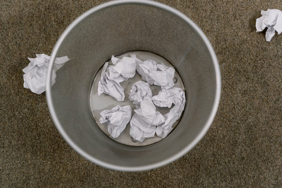 The image shows a cylindrical metal rubbish bin placed on a beige or light brown carpeted floor, viewed from above. Inside the bin, there are several crumpled sheets of white paper, some partially submerged in a small amount of water at the bottom of the bin. A few crumpled paper balls are scattered around the exterior of the bin, resting on the floor. The metallic surface of the bin appears smooth with a slight sheen, and the crumpled papers exhibit varying textures, from tightly wrinkled to loosely crumpled. The environment suggests an indoor setting, possibly an office or domestic space, with the focus on waste disposal or on-site rubbish collection. The scene emphasizes the concept of waste management, relevant to private waste handling and rubbish removal services offered by Rubbish Removal Dulwich, supporting the context of alternative rubbish collection methods such as on-site clearance rather than standard public disposal systems.