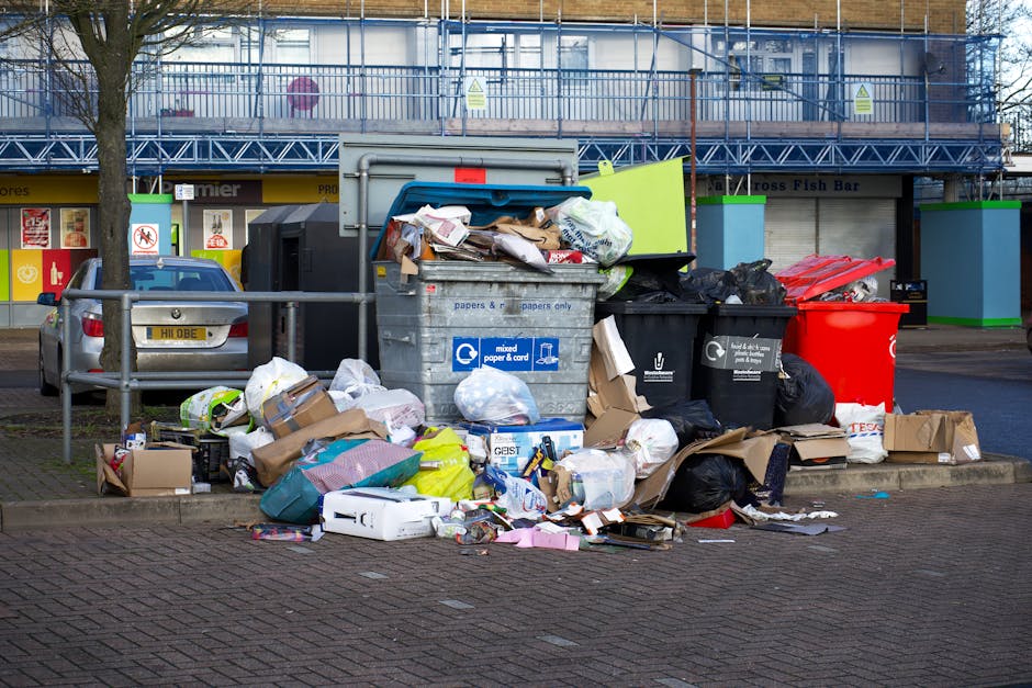 A large pile of mixed rubbish and waste materials displayed on a paved area in front of a small shopping centre, consisting of overflowing black trash bags, flattened cardboard boxes, crumpled paper, plastic packaging, and discarded packaging material scattered across the ground. The waste is positioned beside a metal barrier, partially blocking a parked silver hatchback car with a yellow license plate, and adjacent to multiple recycling bins in various colours including black, red, and black with a recycling symbol, indicating different waste categories. Behind the waste, there is a building with a blue scaffolding structure and storefronts with signage, one of which reads 'Cross Fish Bar,' suggesting an urban environment. The scene appears to be an informal rubbish accumulation, possibly related to private waste collection, and highlights the need for professional rubbish removal services like those offered by Rubbish Removal Dulwich to manage such waste effectively and maintain cleanliness.