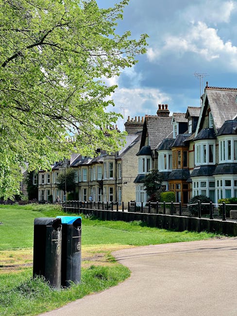 A row of traditional terraced houses with bay windows and tiled roofs behind a low black metal fence, situated in a residential area with a paved footpath in the foreground. To the left, a large tree with fresh green leaves extends its branches over the scene, partially shading the grass and pavement. In the foreground, two black waste bins with blue lids are placed near the pathway, indicating a domestic waste collection point. The scene is set outdoors on a bright day with partly cloudy sky, natural light illuminating the surroundings, and the overall appearance suggesting a typical urban neighbourhood. The presence of waste bins aligns with private rubbish collection services, such as those provided by Rubbish Removal Dulwich, supporting alternative waste management options for local residents.